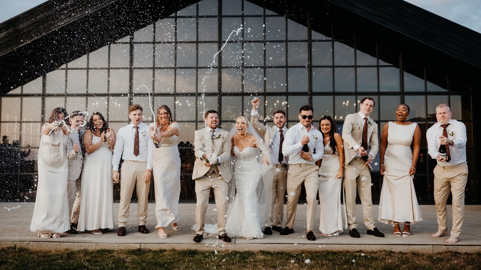 A group of wedding guests, including the bride and groom, stand in a line on a paved area outside a