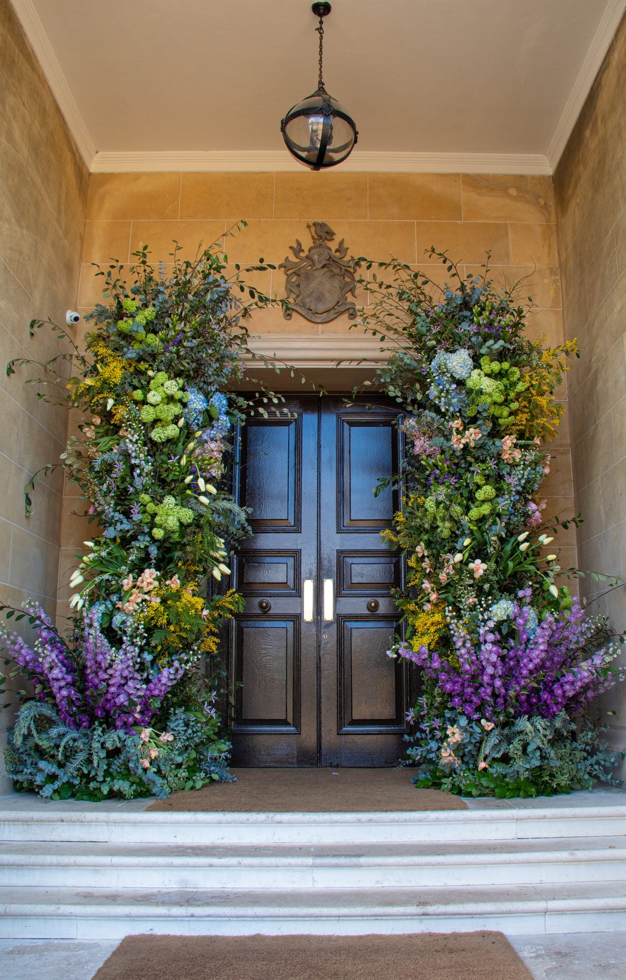 A grand floral installation frames a dark wooden double door at the entrance of a stone building