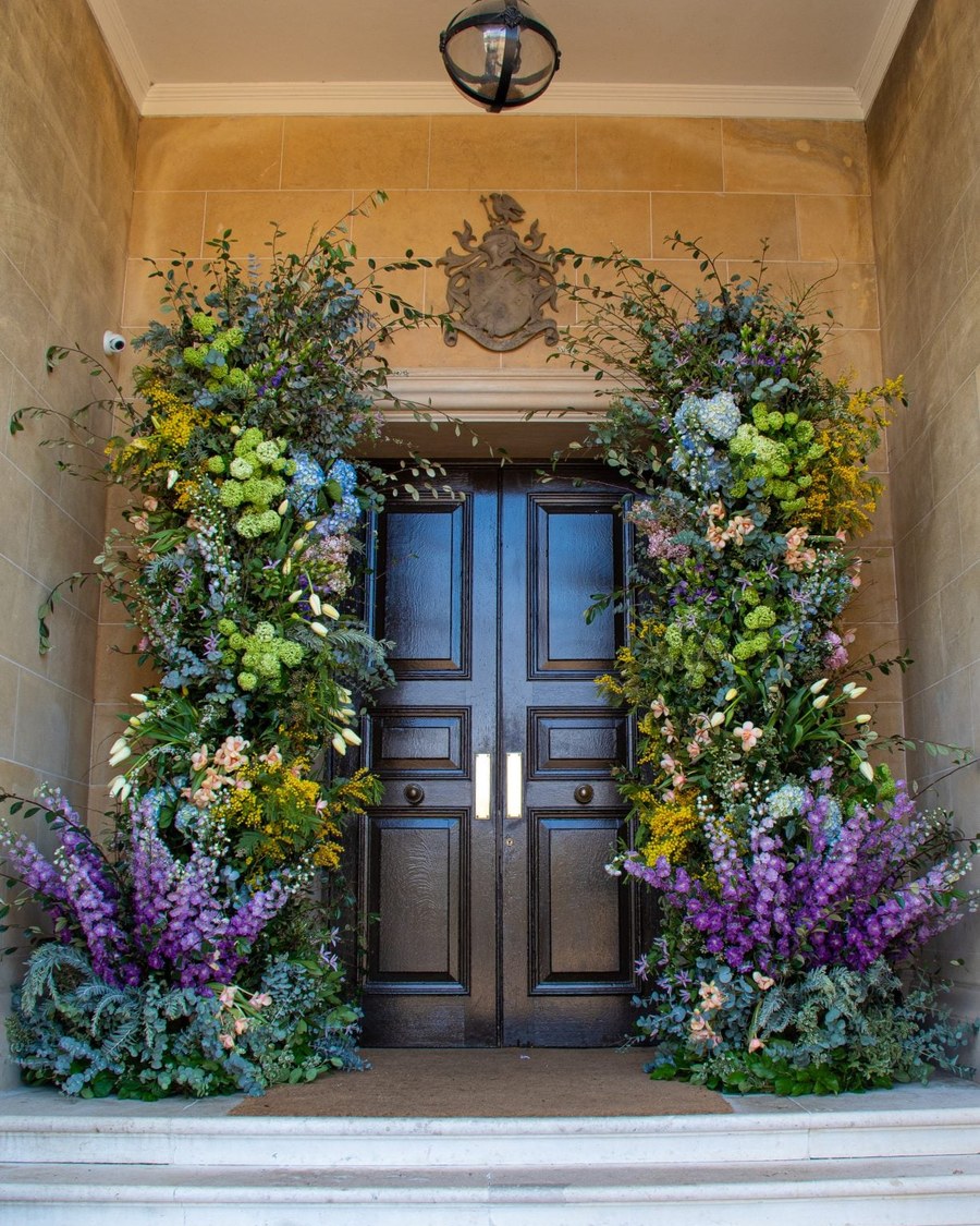 A grand floral installation frames a dark wooden double door at the entrance of a stone building