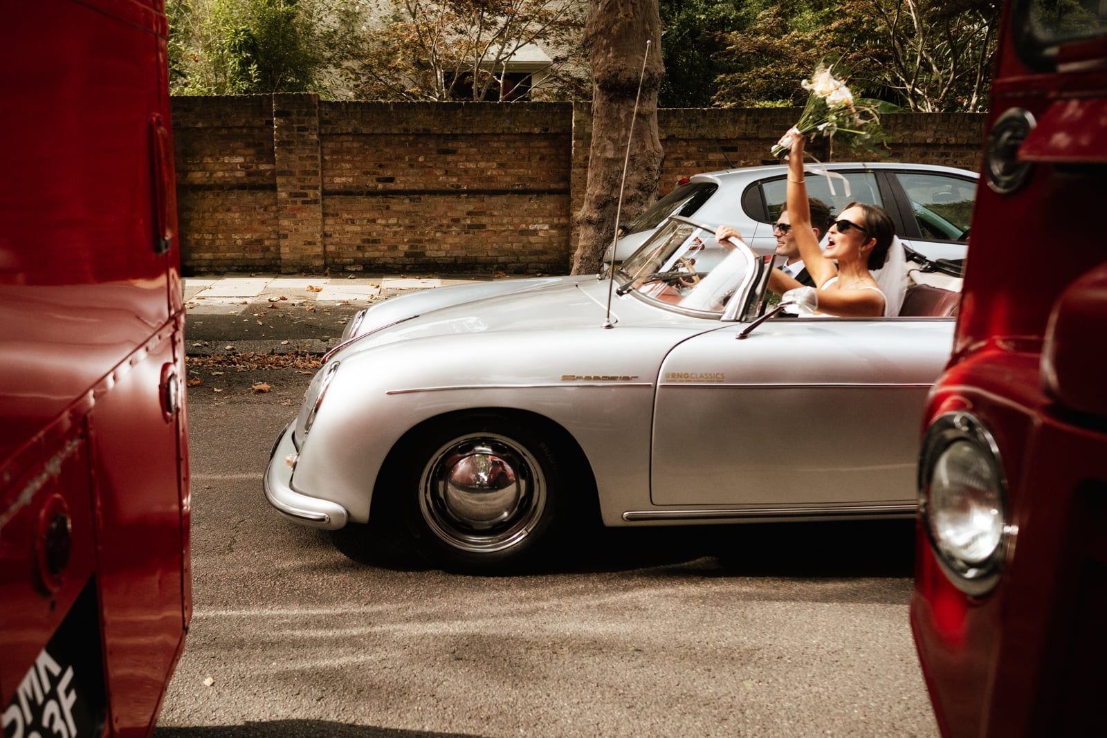 A newlywed couple in a silver classic car navigates a street, with the bride raising her bouquet in