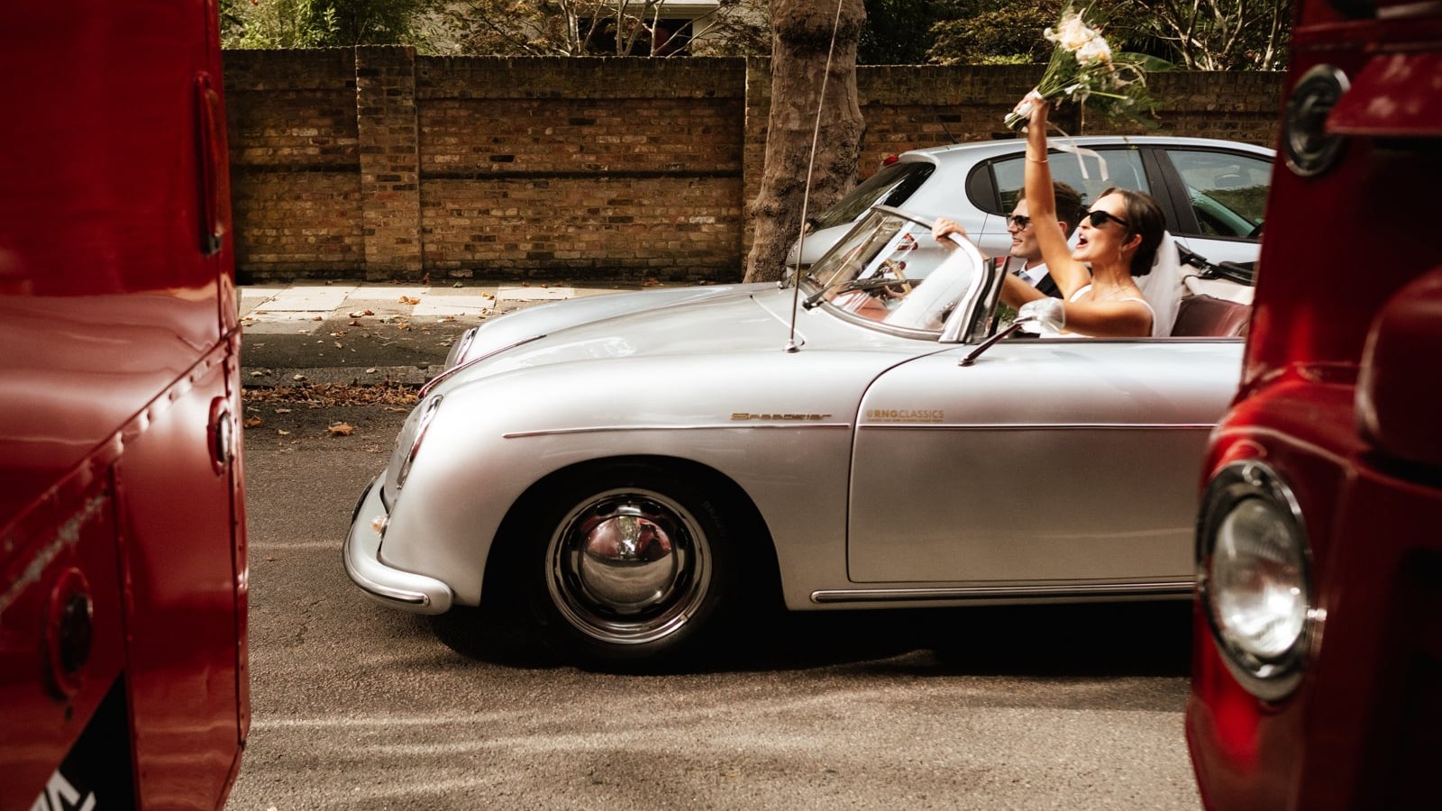 A newlywed couple in a silver classic car navigates a street, with the bride raising her bouquet in