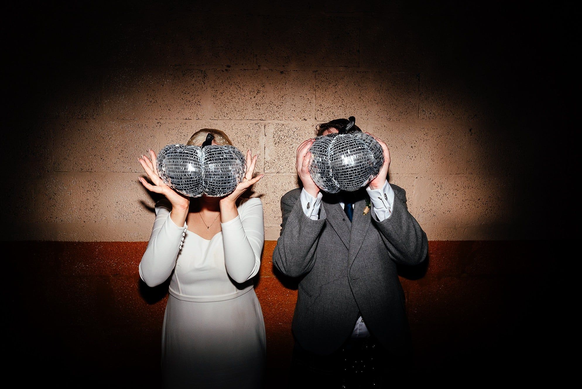 A couple is captured holding small disco ball pumpkins in front of their faces, standing against a