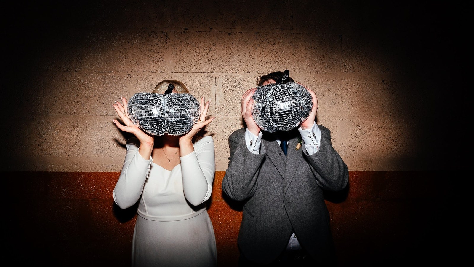 A couple is captured holding small disco ball pumpkins in front of their faces, standing against a