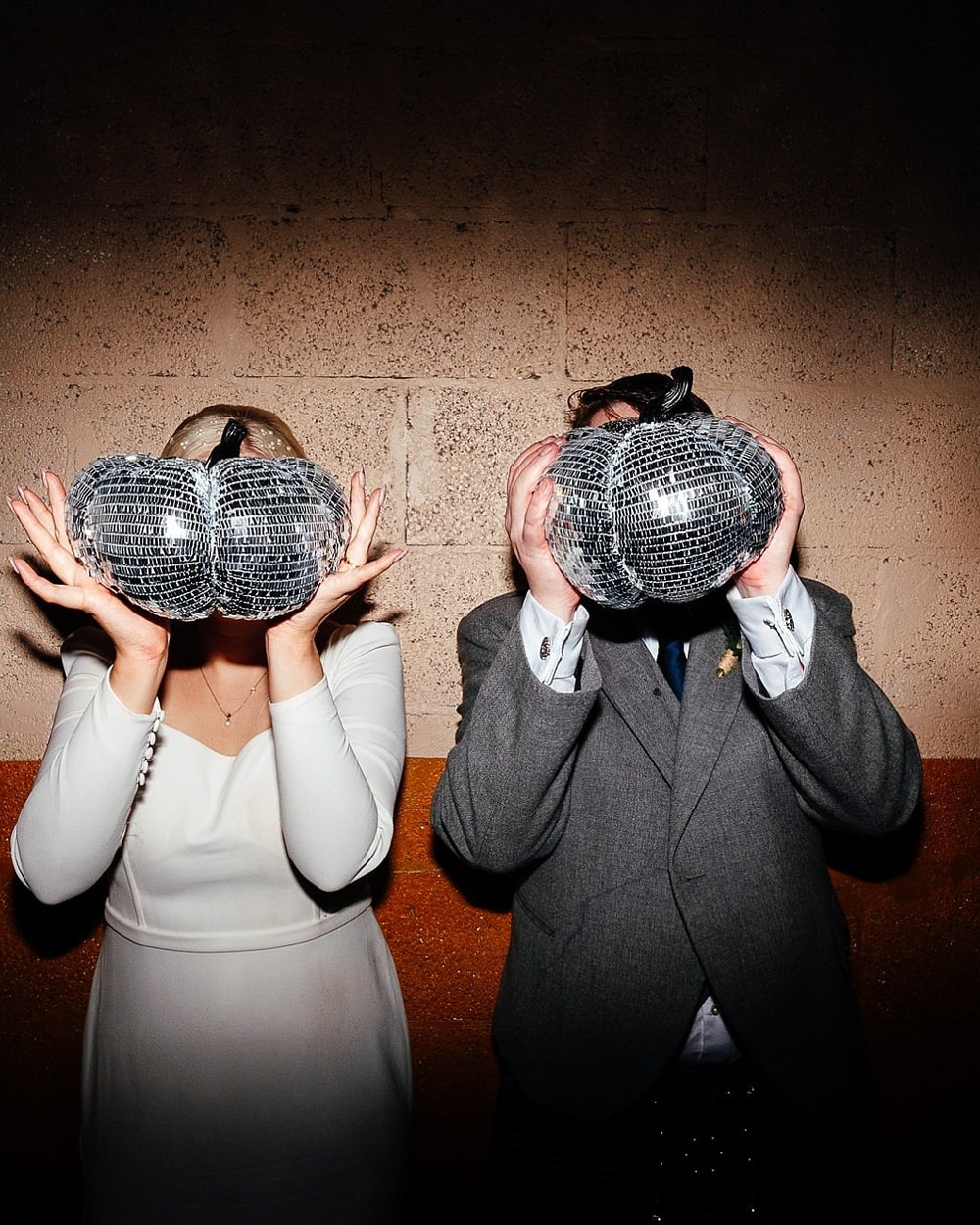 A couple is captured holding small disco ball pumpkins in front of their faces, standing against a