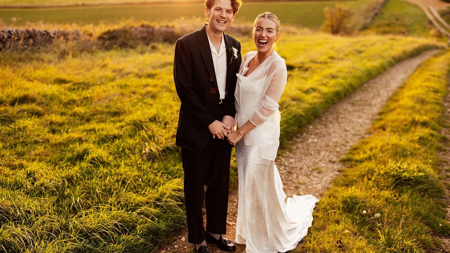 A smiling couple holds hands while standing on a dirt track, surrounded by golden-lit long grass