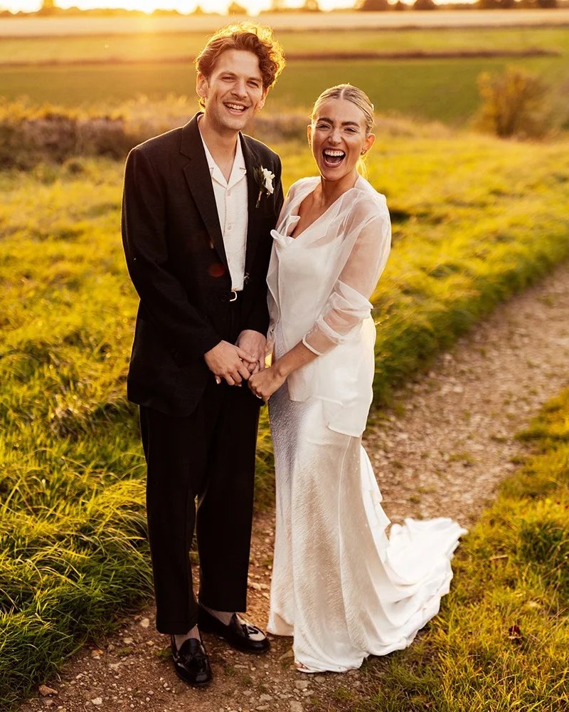 A smiling couple holds hands while standing on a dirt track, surrounded by golden-lit long grass
