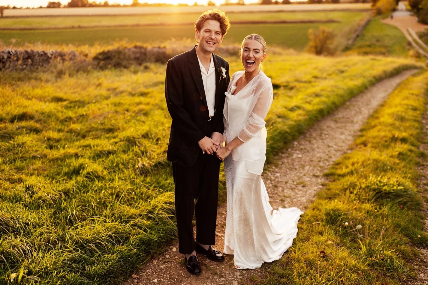A smiling couple holds hands while standing on a dirt track, surrounded by golden-lit long grass