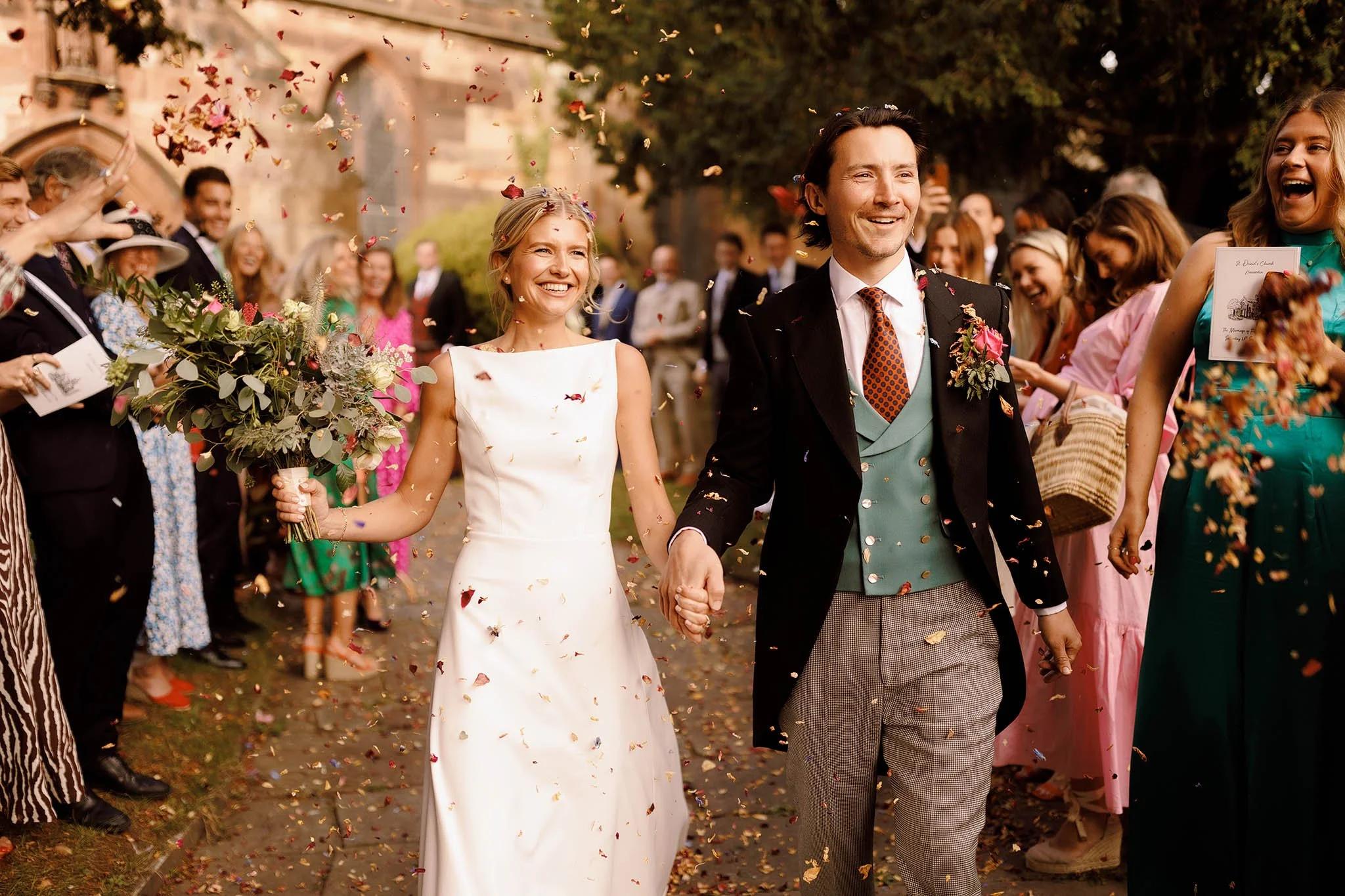 The newly married couple exits a church, holding hands and smiling broadly as guests shower them