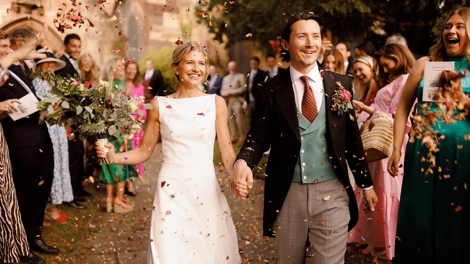 The newly married couple exits a church, holding hands and smiling broadly as guests shower them