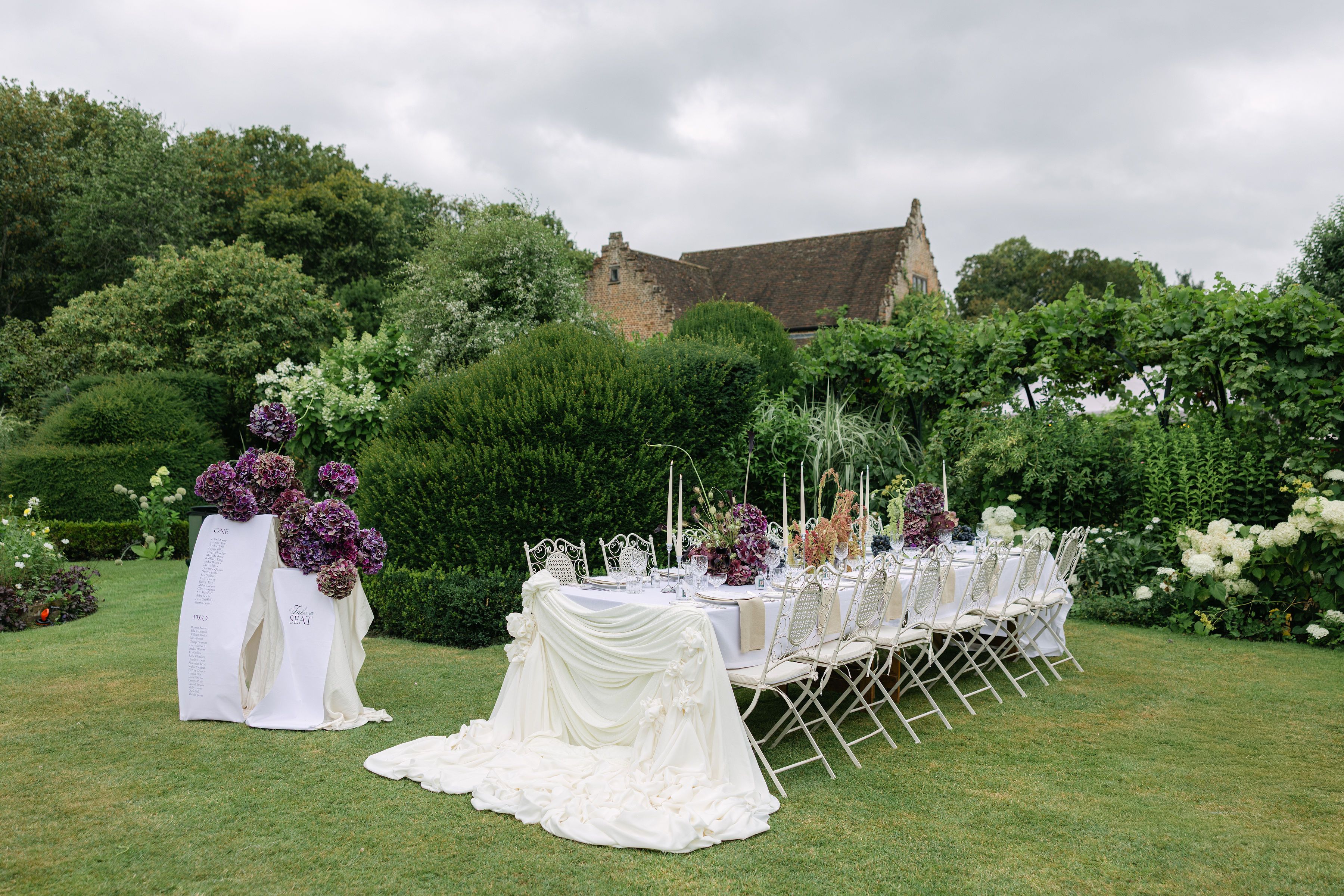 A long outdoor dining table is set on a manicured lawn, draped with white fabric and featuring