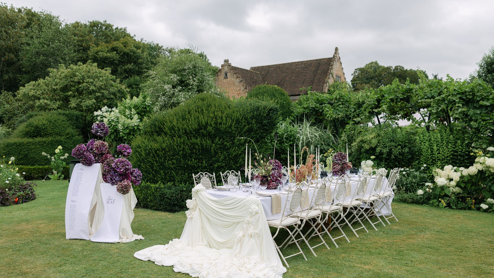 A long outdoor dining table is set on a manicured lawn, draped with white fabric and featuring