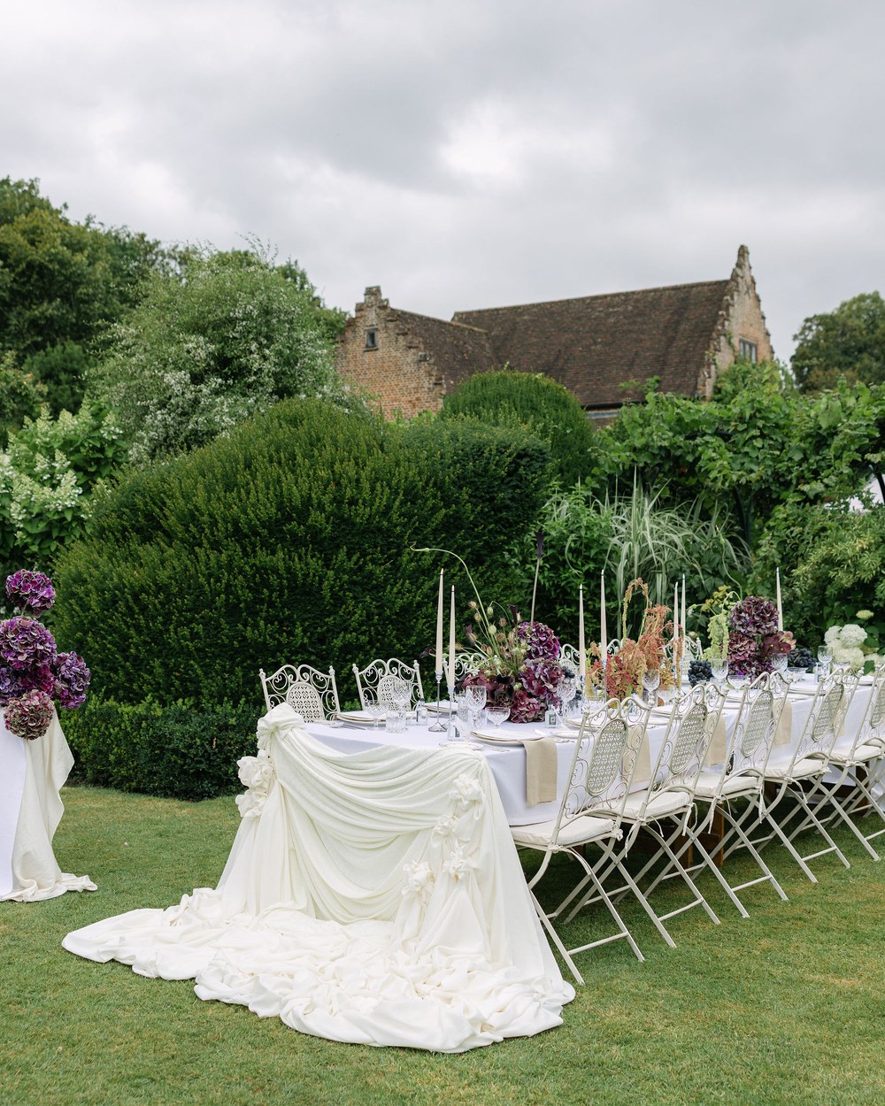 A long outdoor dining table is set on a manicured lawn, draped with white fabric and featuring