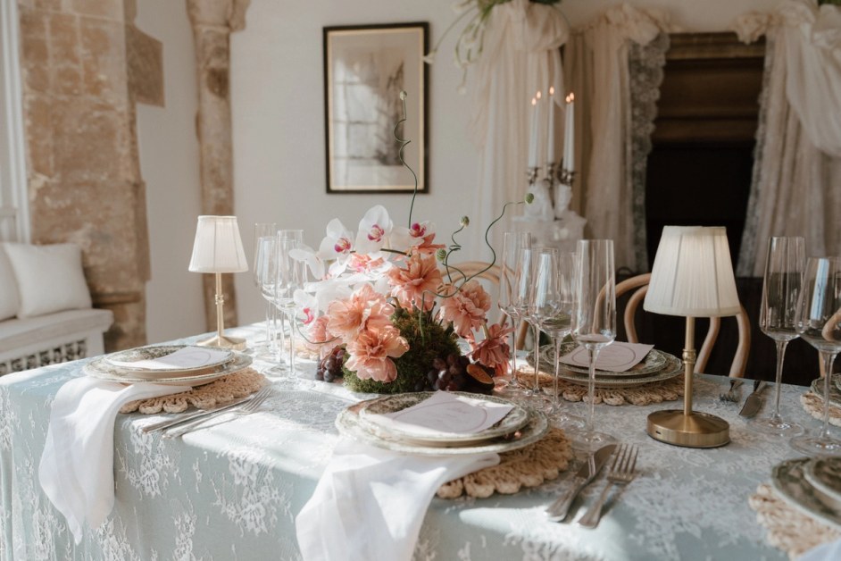 A wedding breakfast table laid with a pale lace cloth, crochet placemats and small gold lamps, centred on a low arrangement of peach carnations, white orchids and grapes in a stone room.