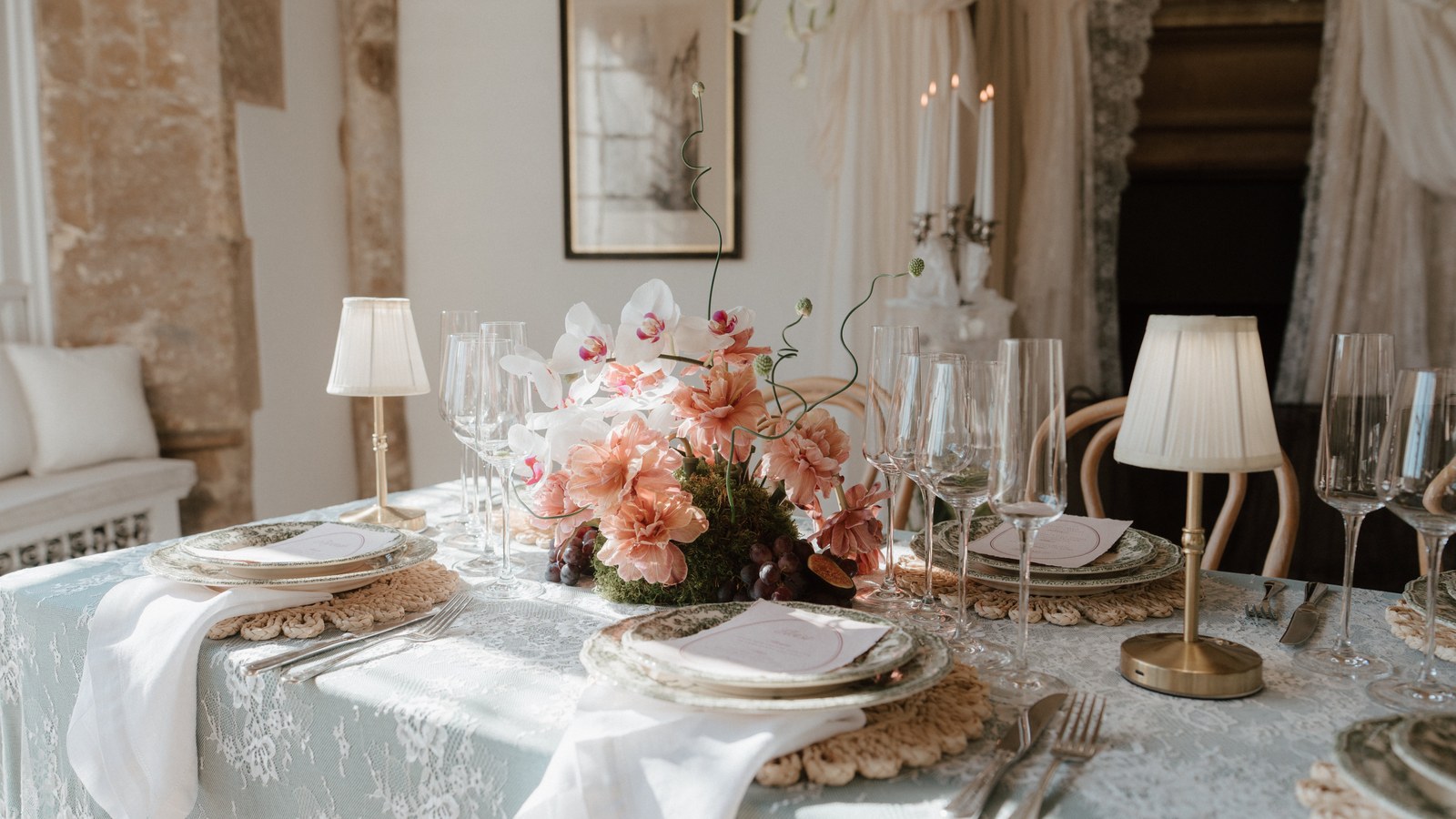 A wedding breakfast table laid with a pale lace cloth, crochet placemats and small gold lamps, centred on a low arrangement of peach carnations, white orchids and grapes in a stone room.