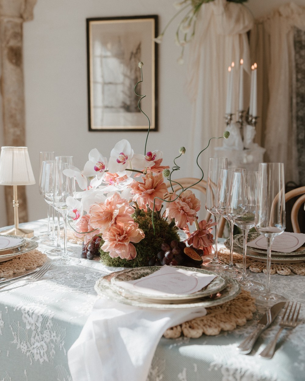A wedding breakfast table laid with a pale lace cloth, crochet placemats and small gold lamps, centred on a low arrangement of peach carnations, white orchids and grapes in a stone room.