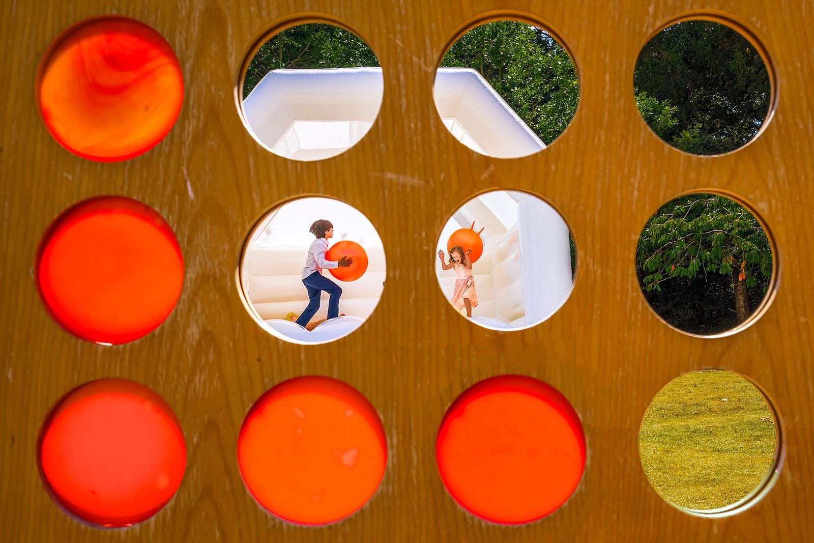 Children play on an inflatable bouncy castle, viewed through circular cut-outs in a wooden board.