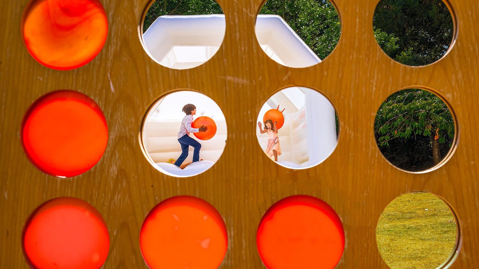 Children play on an inflatable bouncy castle, viewed through circular cut-outs in a wooden board.