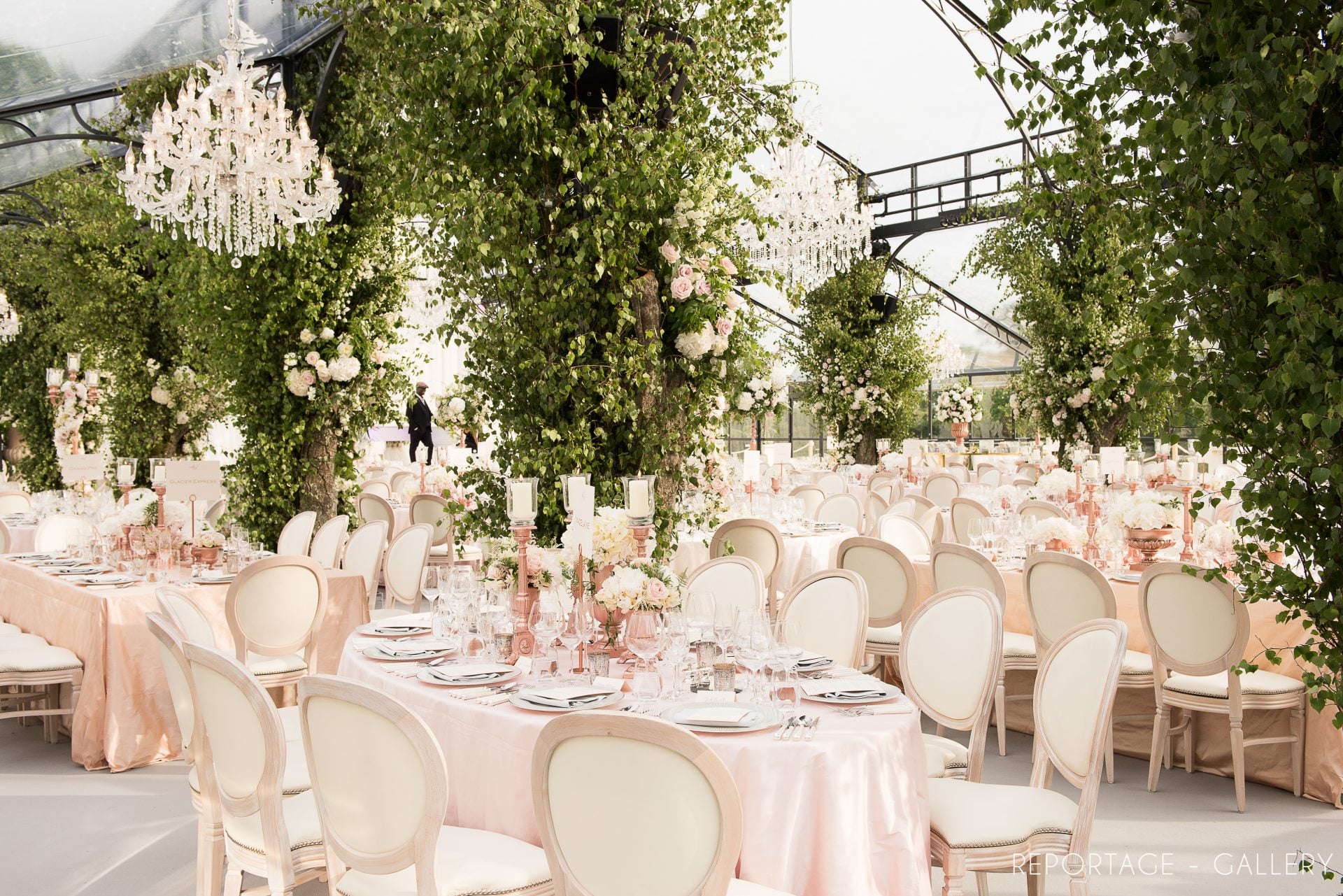A spacious wedding reception setting under a clear marquee, featuring numerous round tables draped