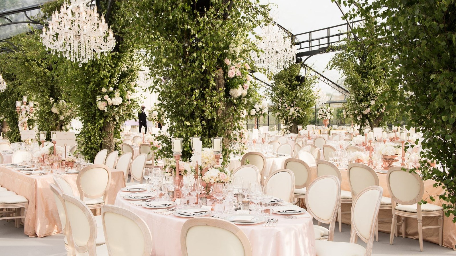 A spacious wedding reception setting under a clear marquee, featuring numerous round tables draped