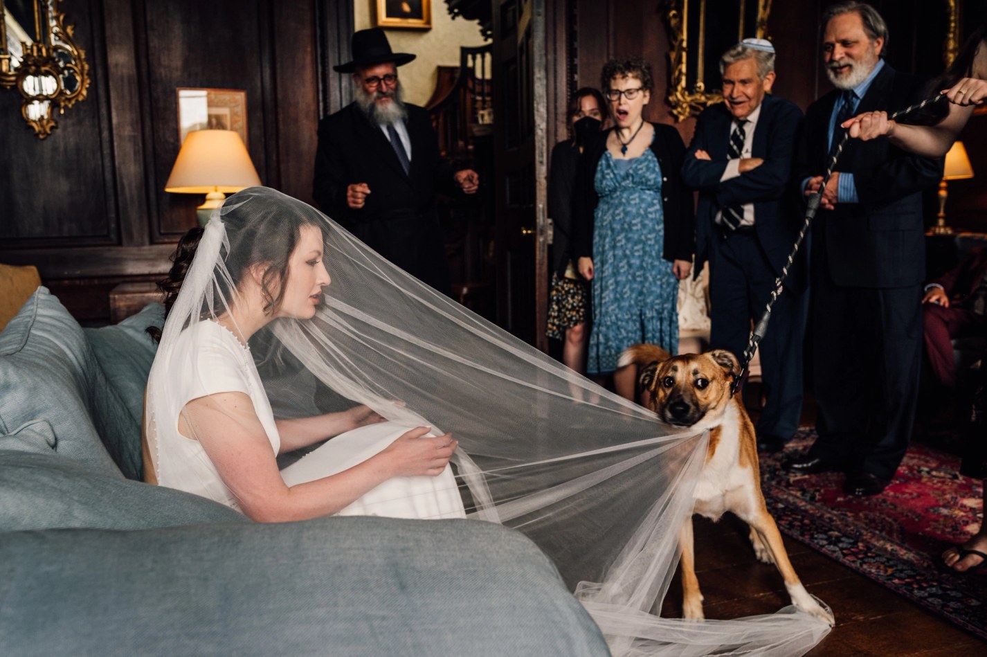 A bride in a short-sleeved white dress and veil sits on a blue-grey sofa, looking towards a light