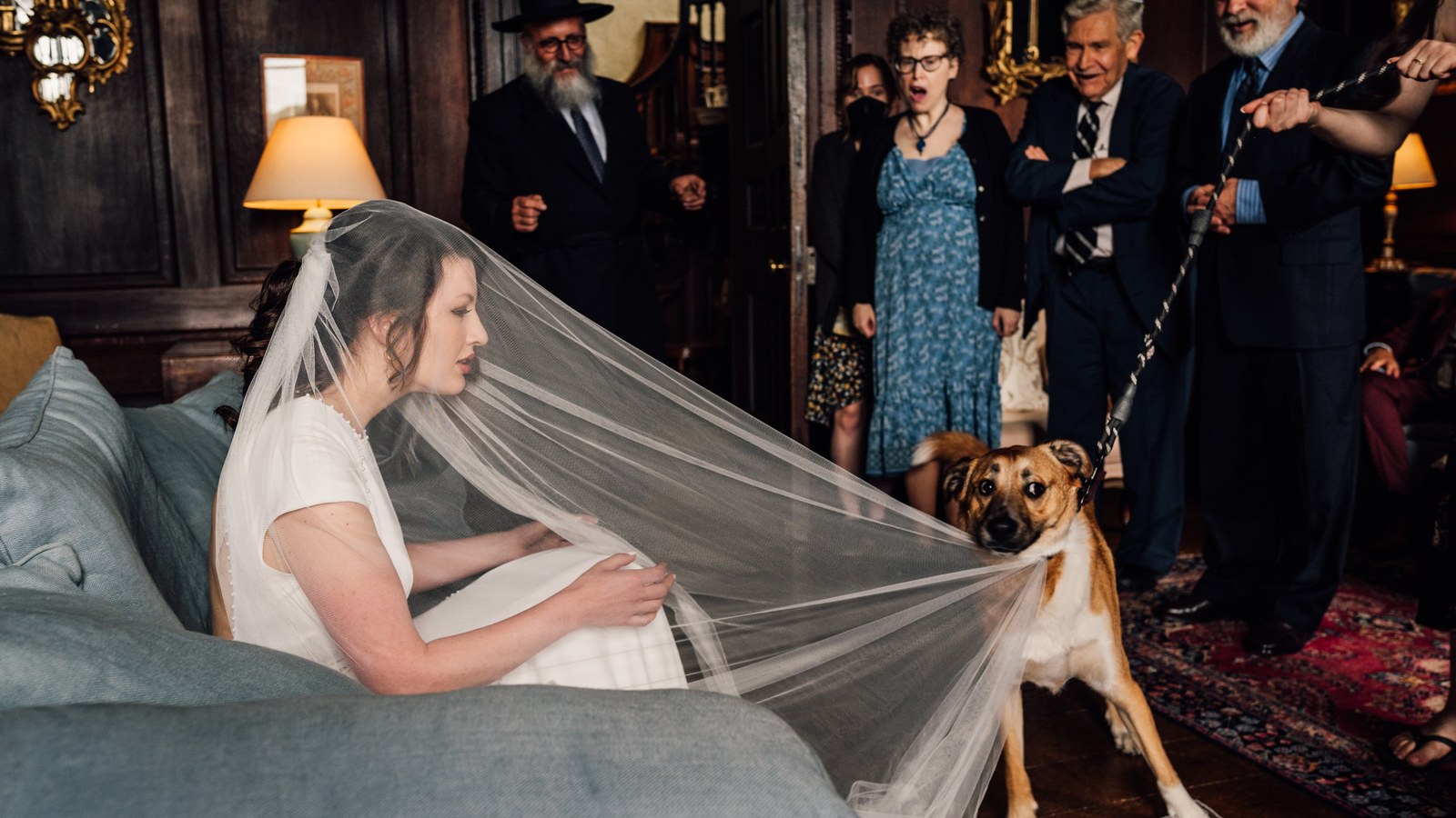 A bride in a short-sleeved white dress and veil sits on a blue-grey sofa, looking towards a light