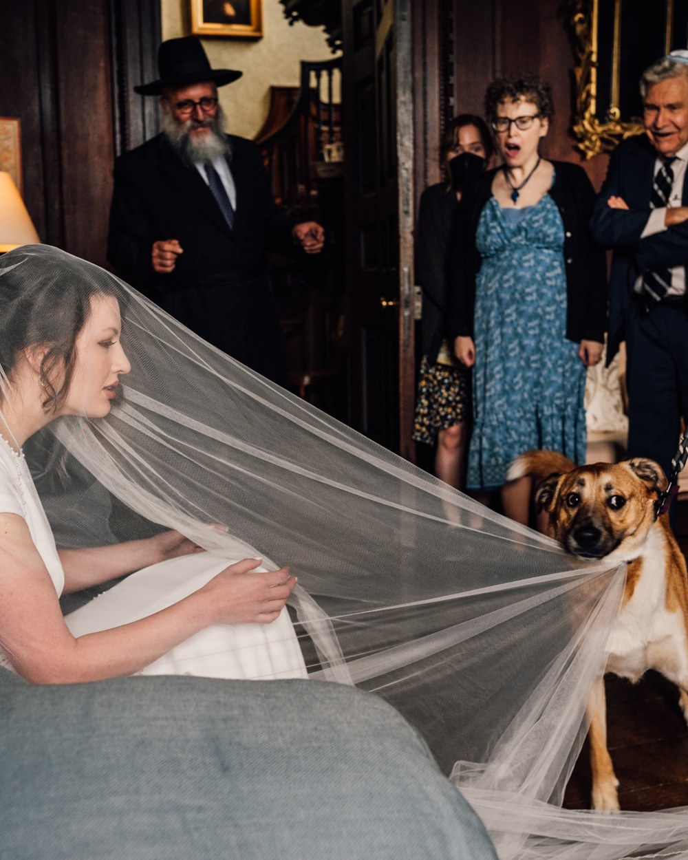 A bride in a short-sleeved white dress and veil sits on a blue-grey sofa, looking towards a light