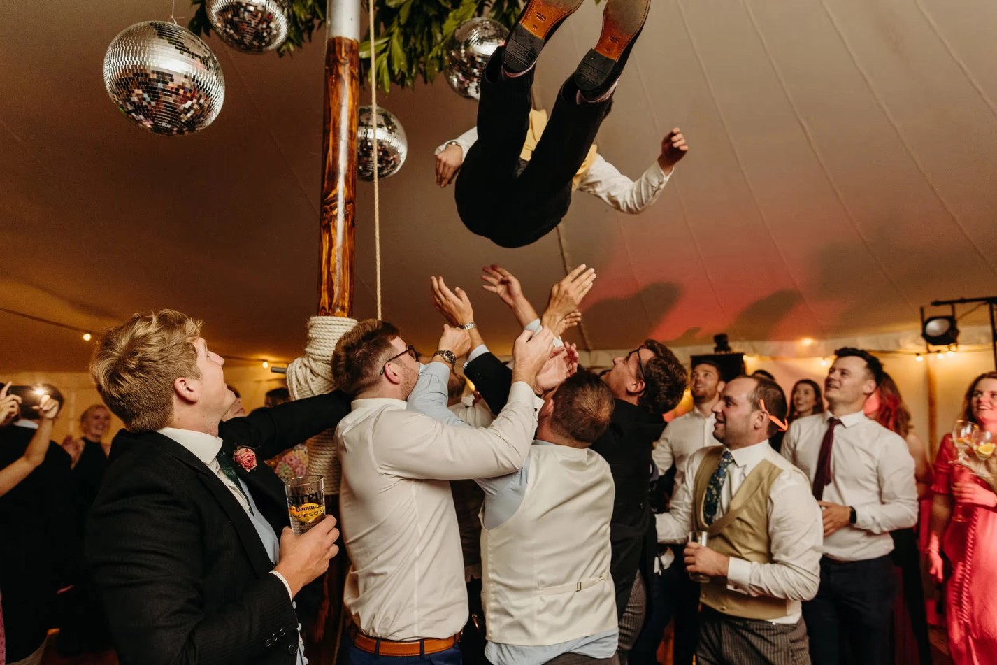 Wedding guests in a marquee toss a groom in the air during the evening reception. The indoor scene