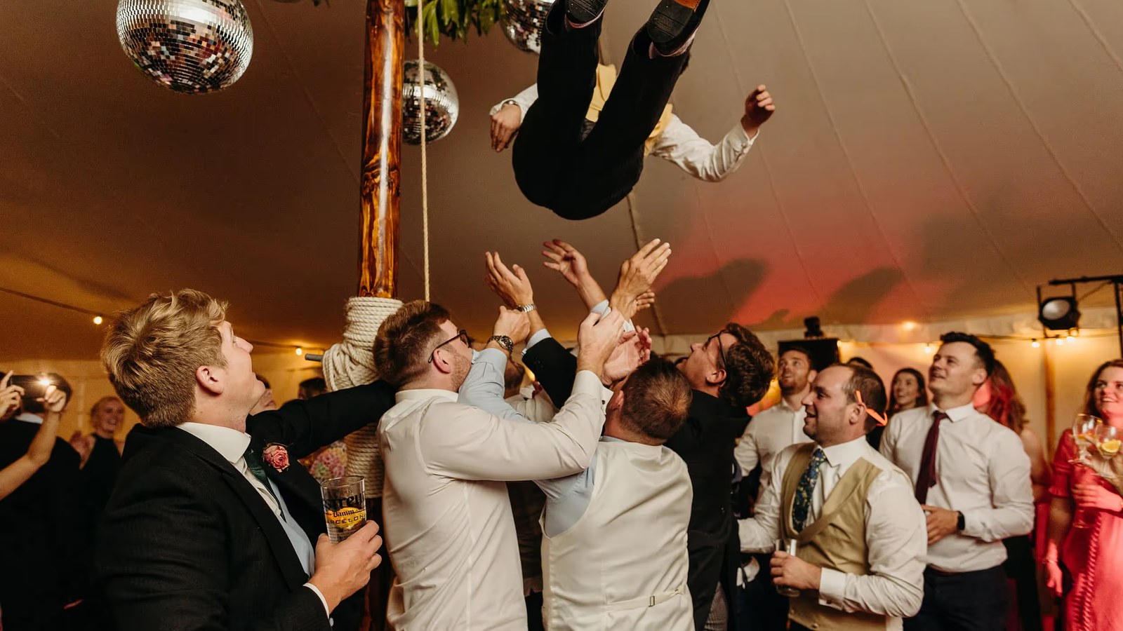 Wedding guests in a marquee toss a groom in the air during the evening reception. The indoor scene