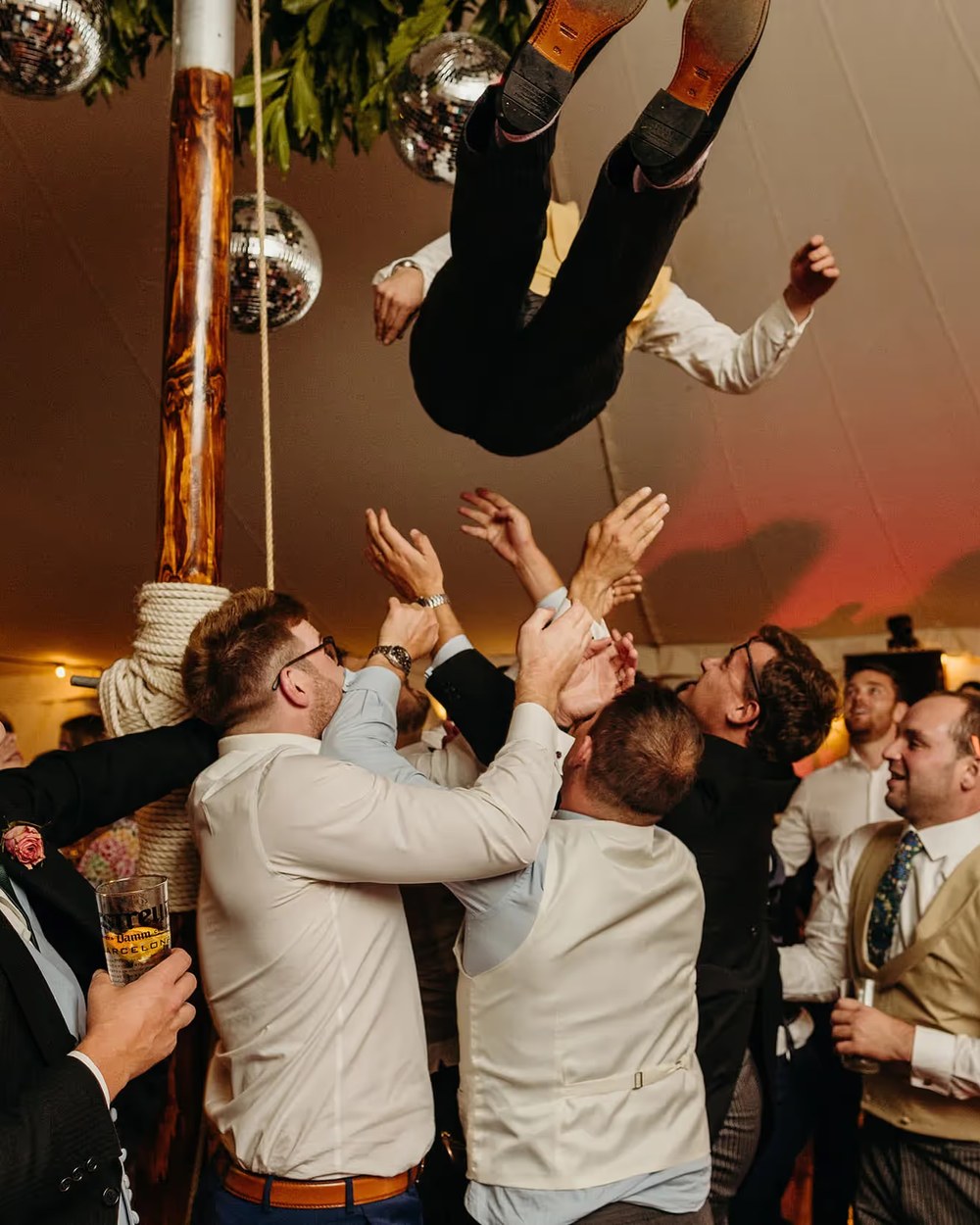 Wedding guests in a marquee toss a groom in the air during the evening reception. The indoor scene