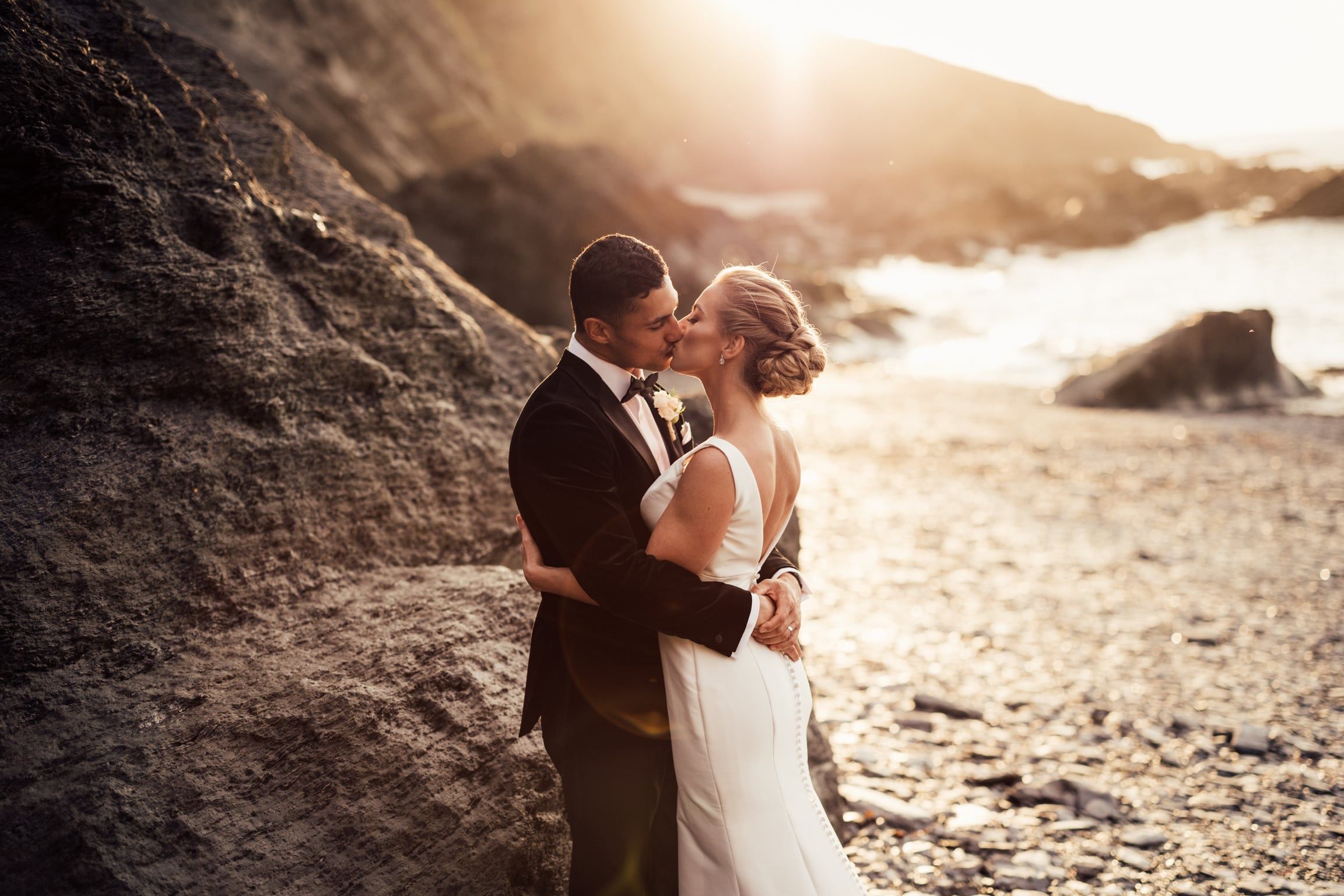 A newlywed couple shares a kiss on a rocky beach at sunset. The bride wears a white backless gown