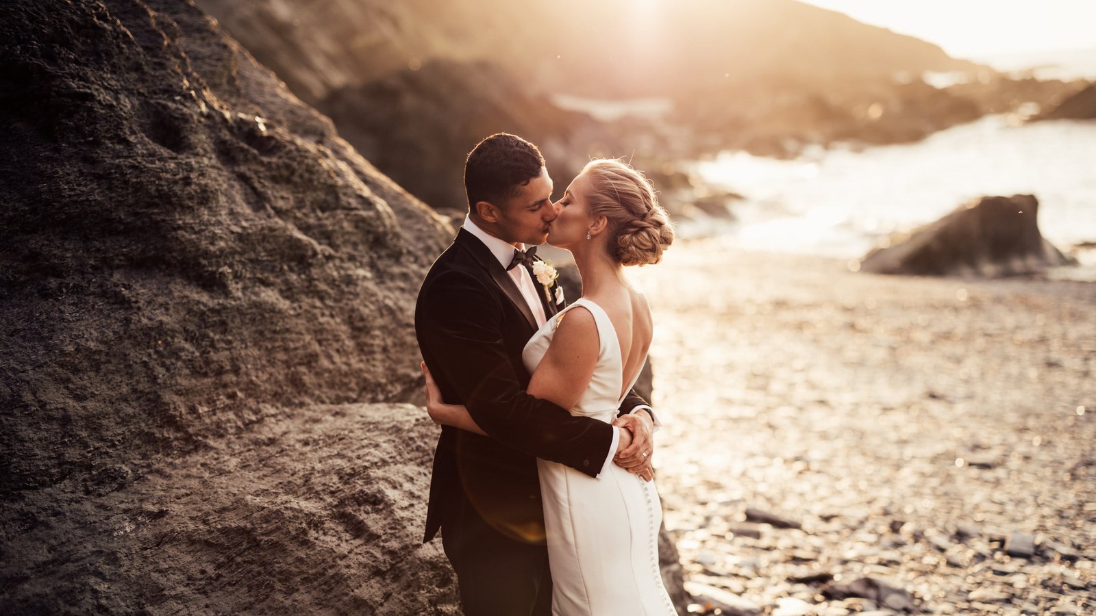 A newlywed couple shares a kiss on a rocky beach at sunset. The bride wears a white backless gown