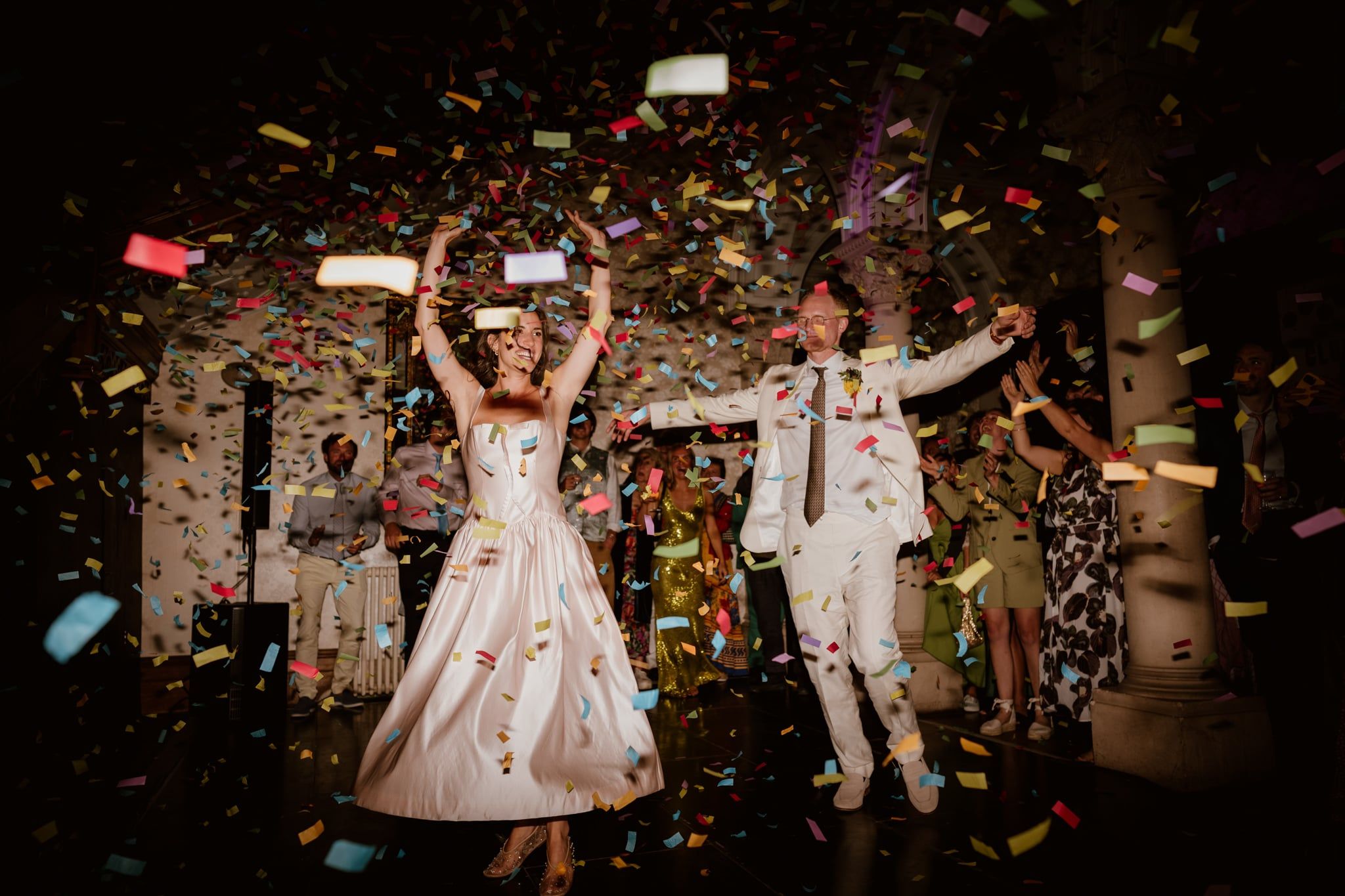 A bride in a pale pink satin dress and a groom in an off-white suit jump and dance amidst falling