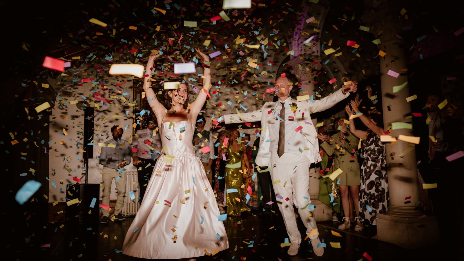 A bride in a pale pink satin dress and a groom in an off-white suit jump and dance amidst falling