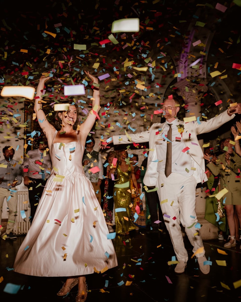 A bride in a pale pink satin dress and a groom in an off-white suit jump and dance amidst falling