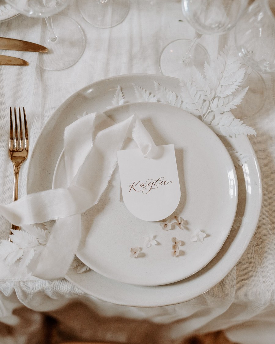 A romantic place card with modern calligraphy, styled on a neutral tablescape. The design features