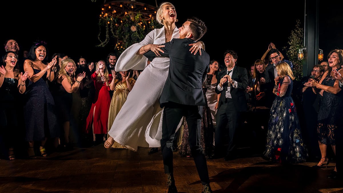 The groom lifts the bride into an exuberant hug on a wooden dance floor, surrounded by cheering and