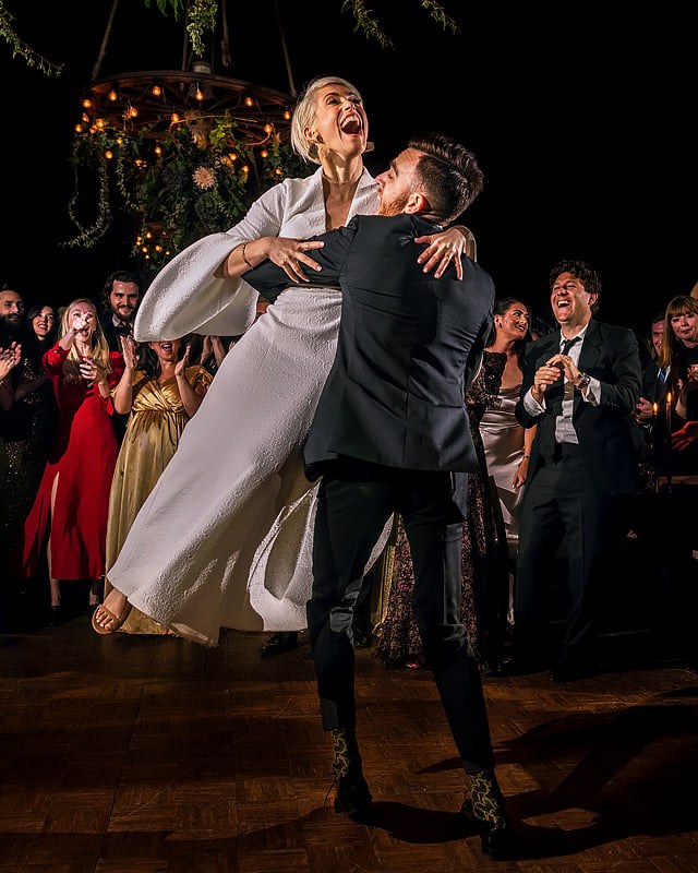 The groom lifts the bride into an exuberant hug on a wooden dance floor, surrounded by cheering and