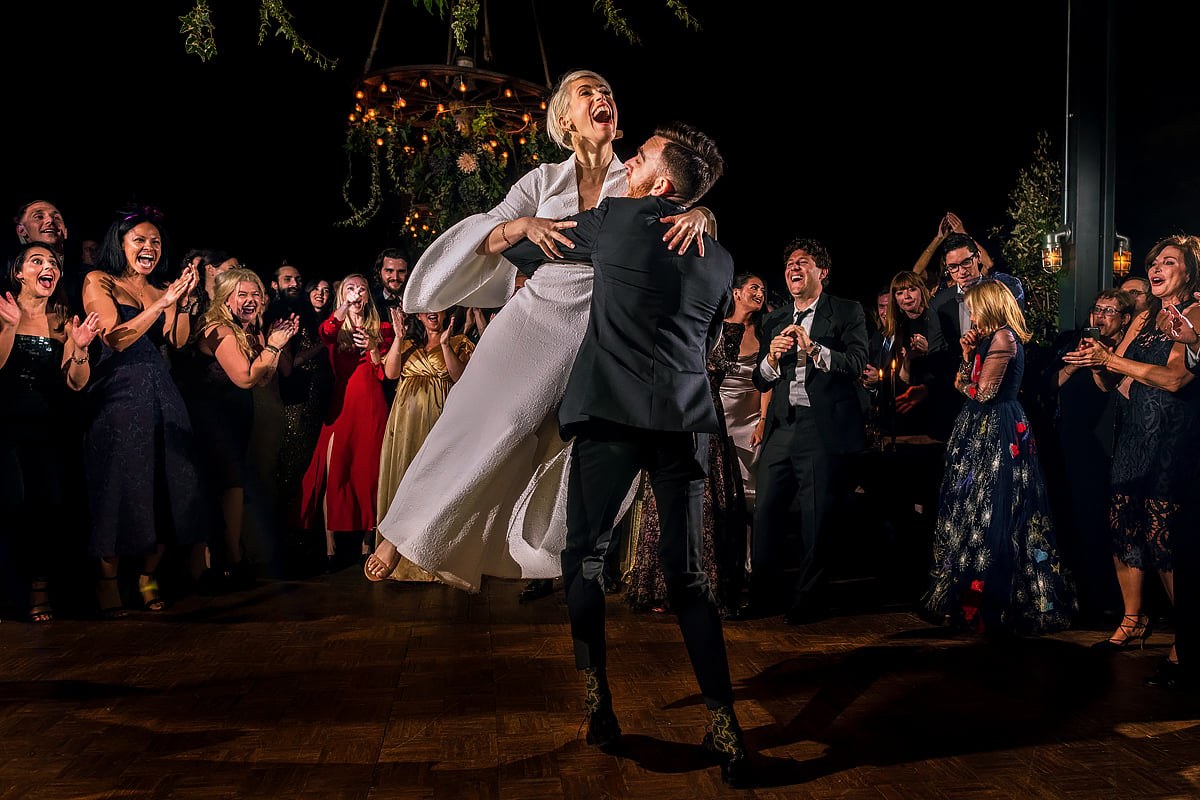 The groom lifts the bride into an exuberant hug on a wooden dance floor, surrounded by cheering and