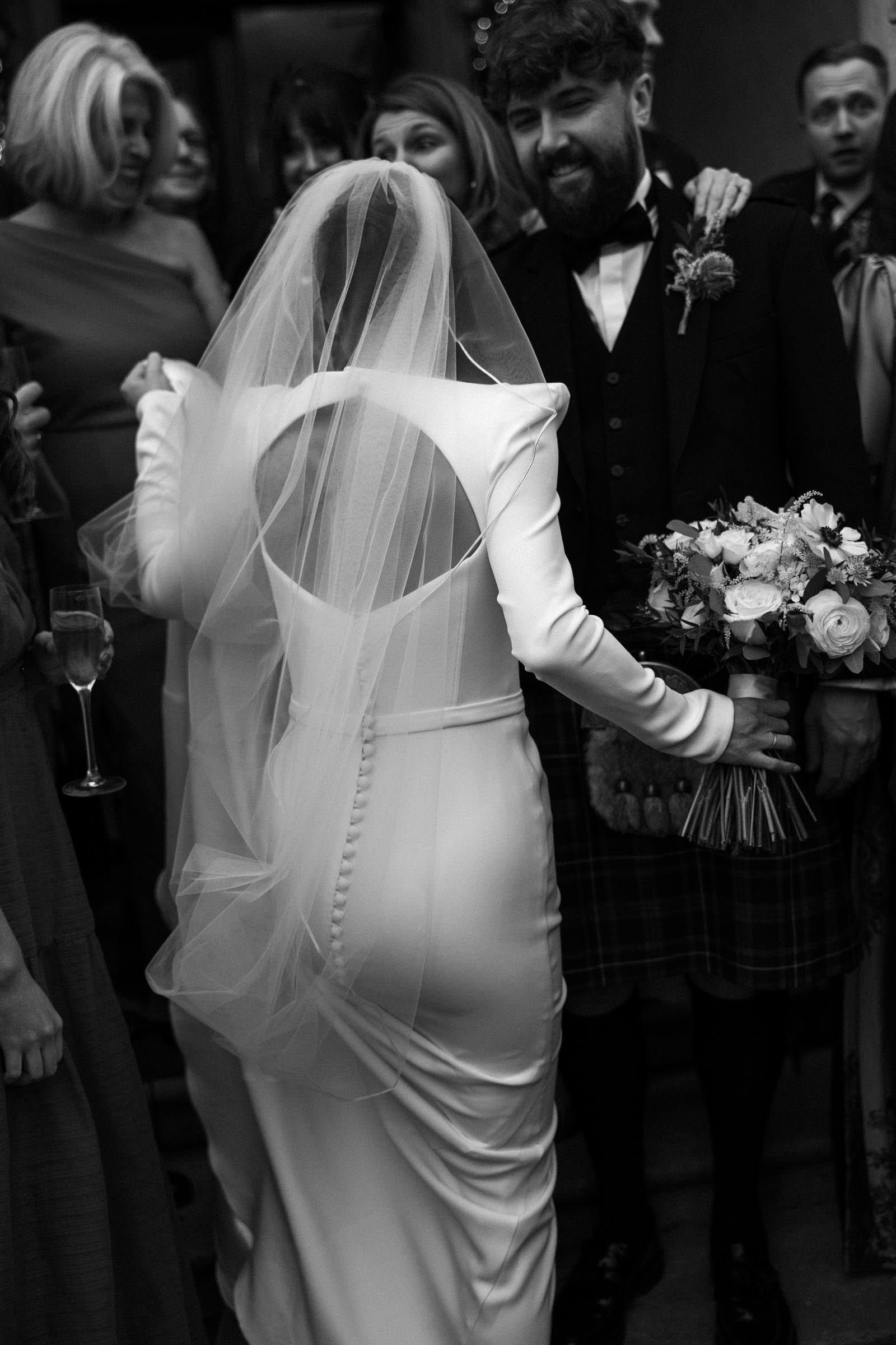 A bride wearing a long-sleeved dress with a large back cutout and a sheer veil stands with her back