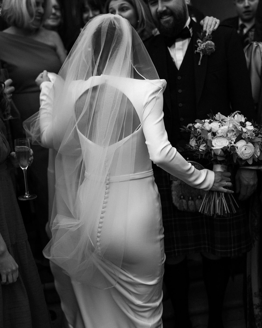 A bride wearing a long-sleeved dress with a large back cutout and a sheer veil stands with her back