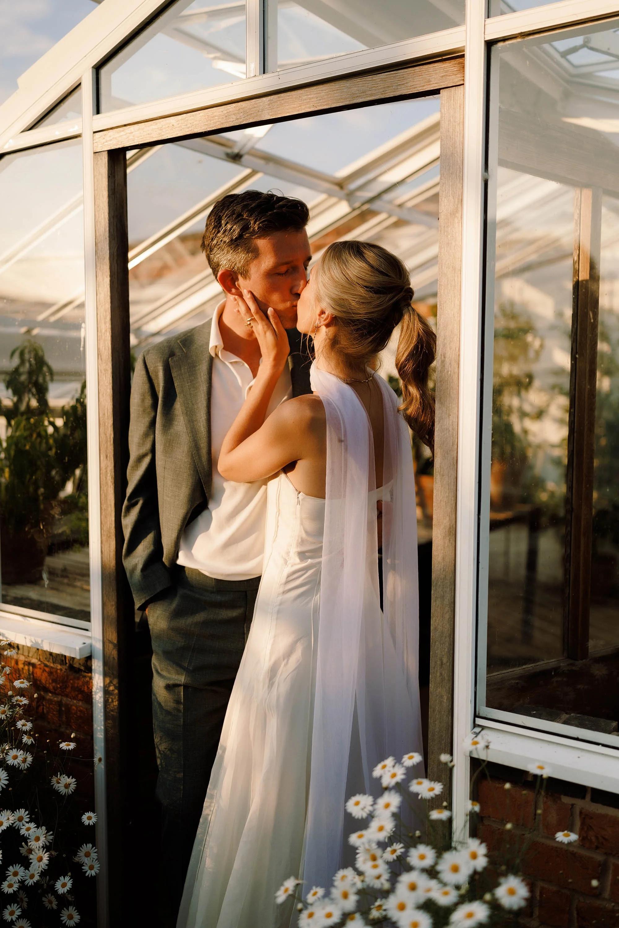 A couple shares a kiss inside a glasshouse, framed by white windowpanes and surrounded by white