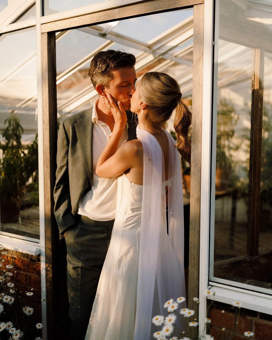 A couple shares a kiss inside a glasshouse, framed by white windowpanes and surrounded by white