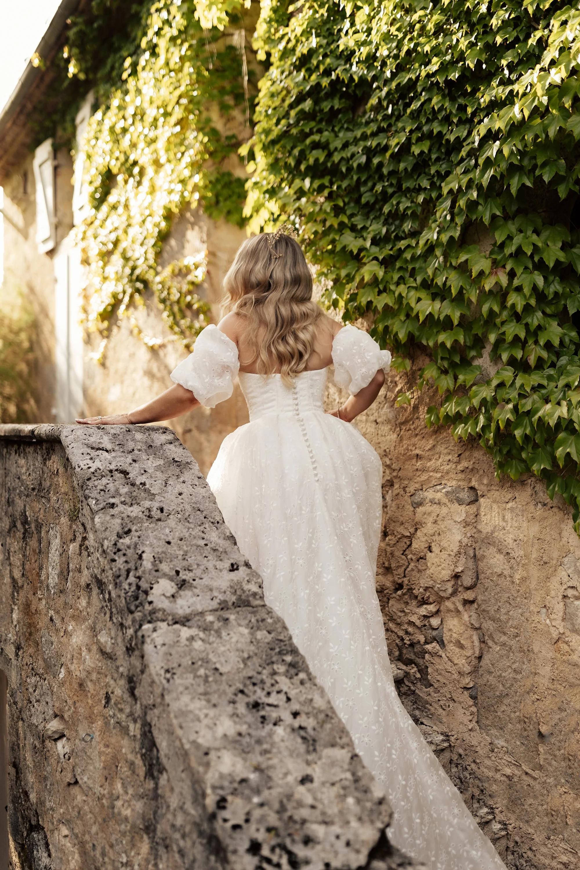 A bride in a white embroidered gown with puff sleeves ascends a stone staircase, with her hand