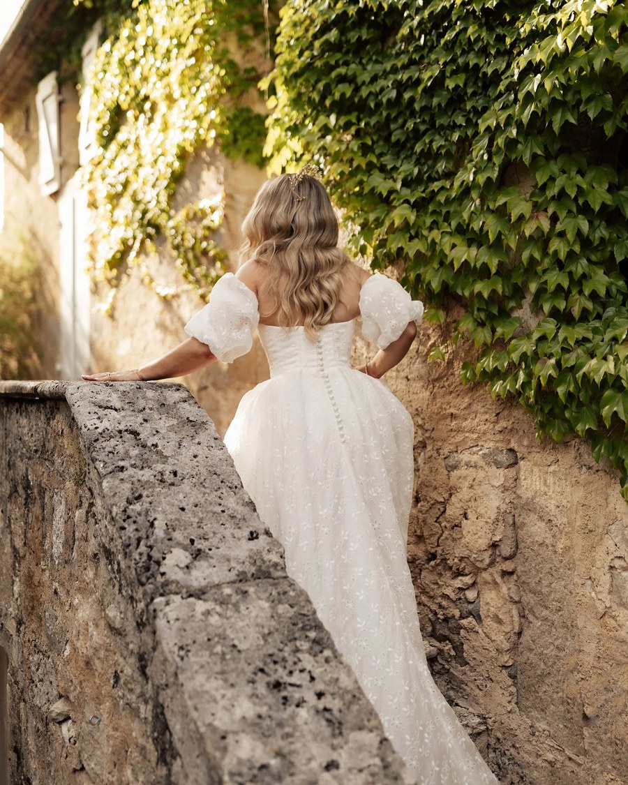 A bride in a white embroidered gown with puff sleeves ascends a stone staircase, with her hand