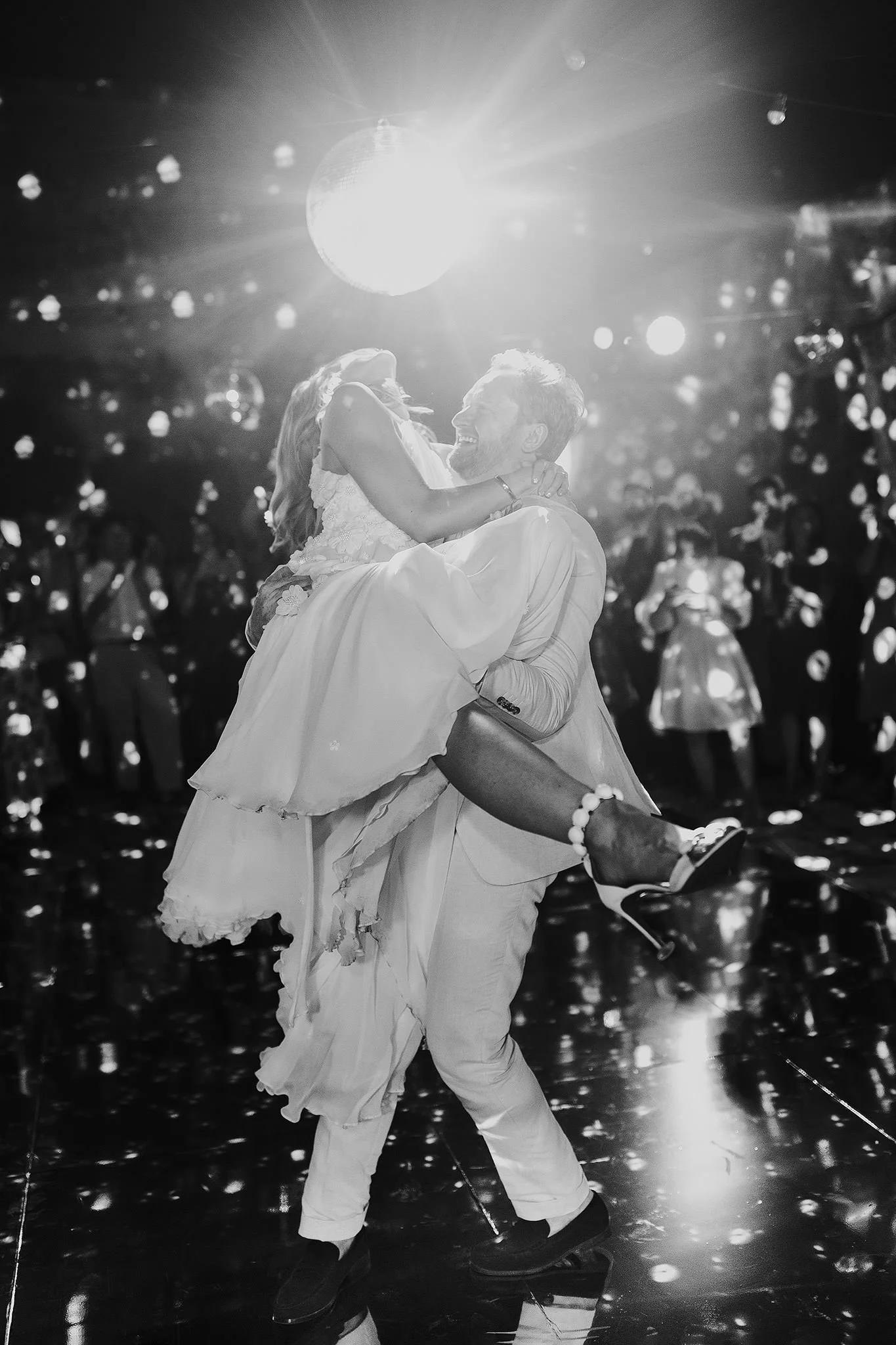 A groom lifts his bride on a reflective dance floor under a bright disco ball, surrounded by guests