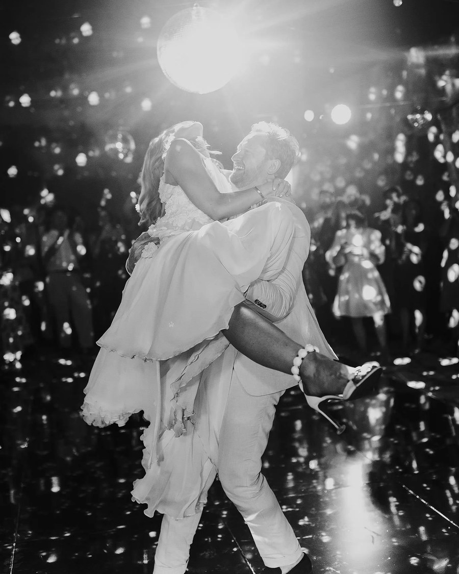 A groom lifts his bride on a reflective dance floor under a bright disco ball, surrounded by guests