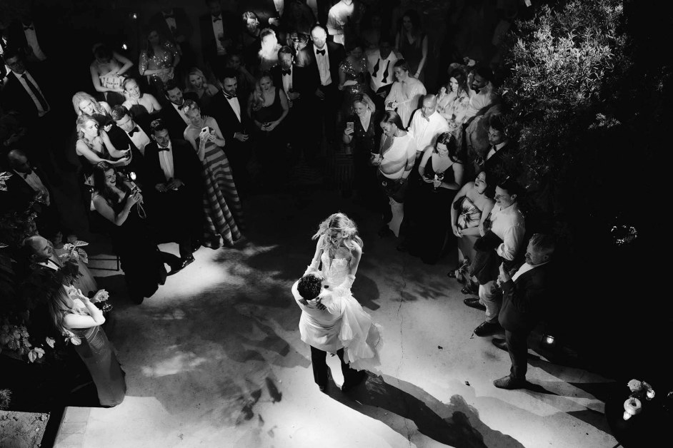 The bride and groom share a first dance in the centre of a reception crowd, illuminated by