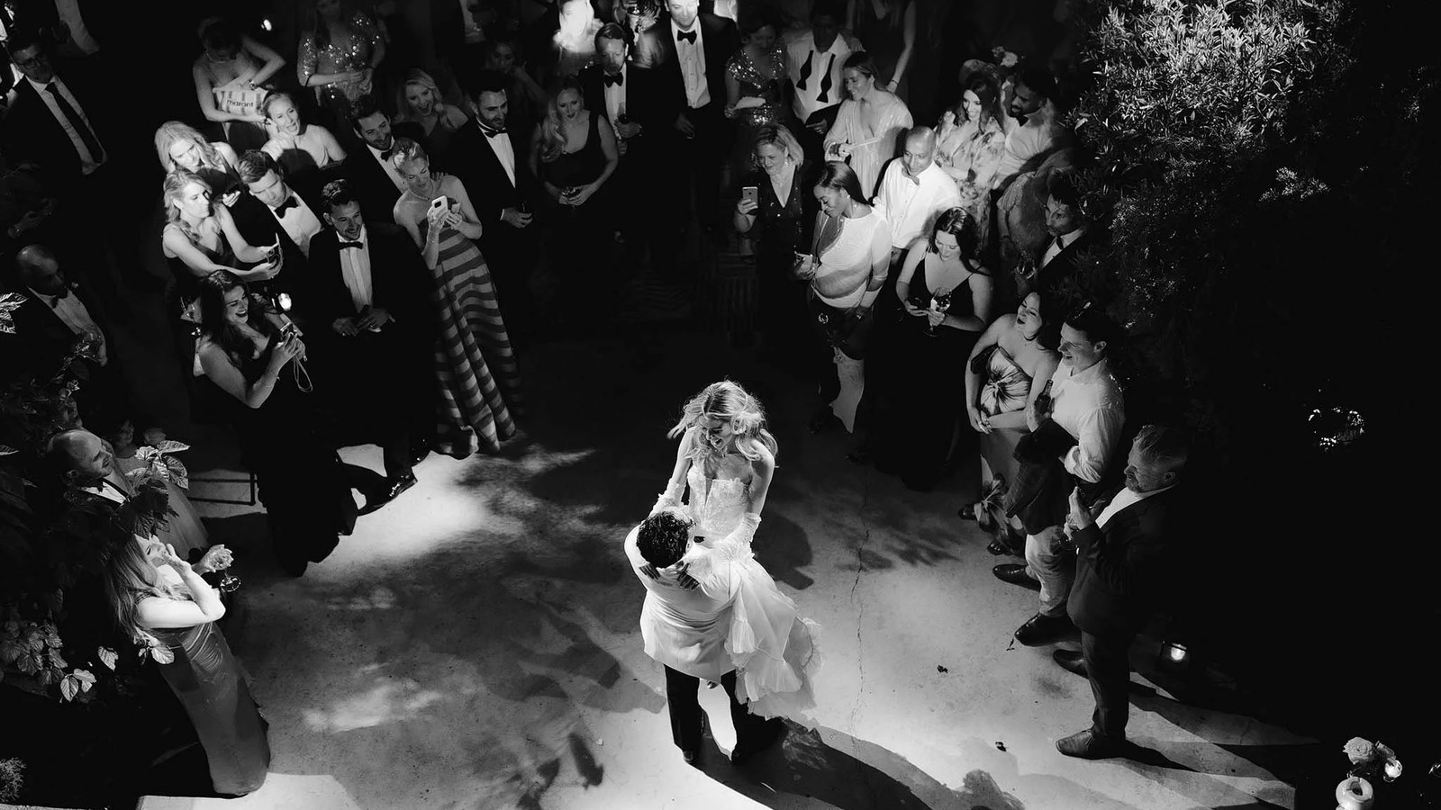 The bride and groom share a first dance in the centre of a reception crowd, illuminated by