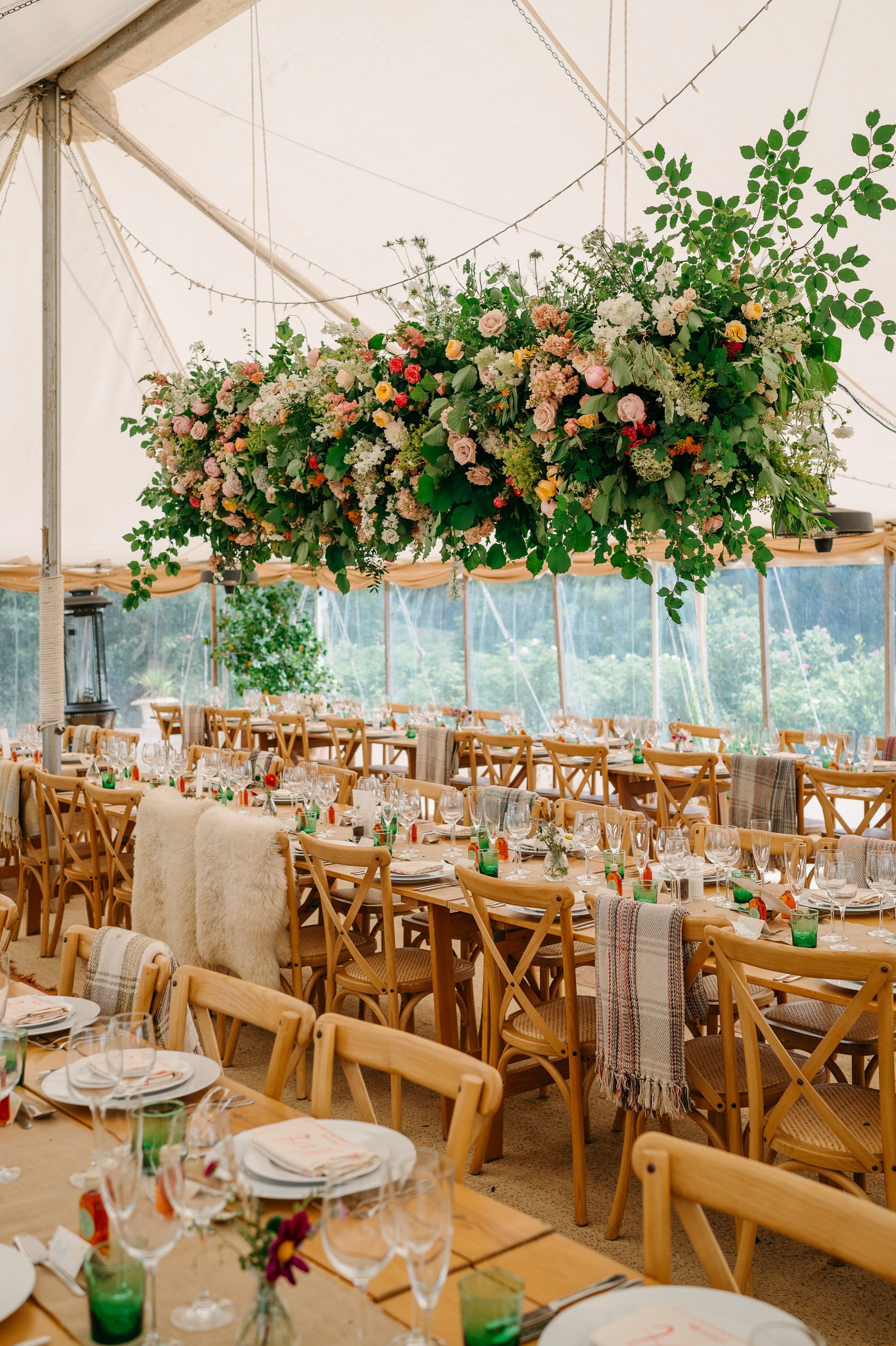 A marquee interior is set with long wooden banquet tables, cross-back chairs, and woven place mats