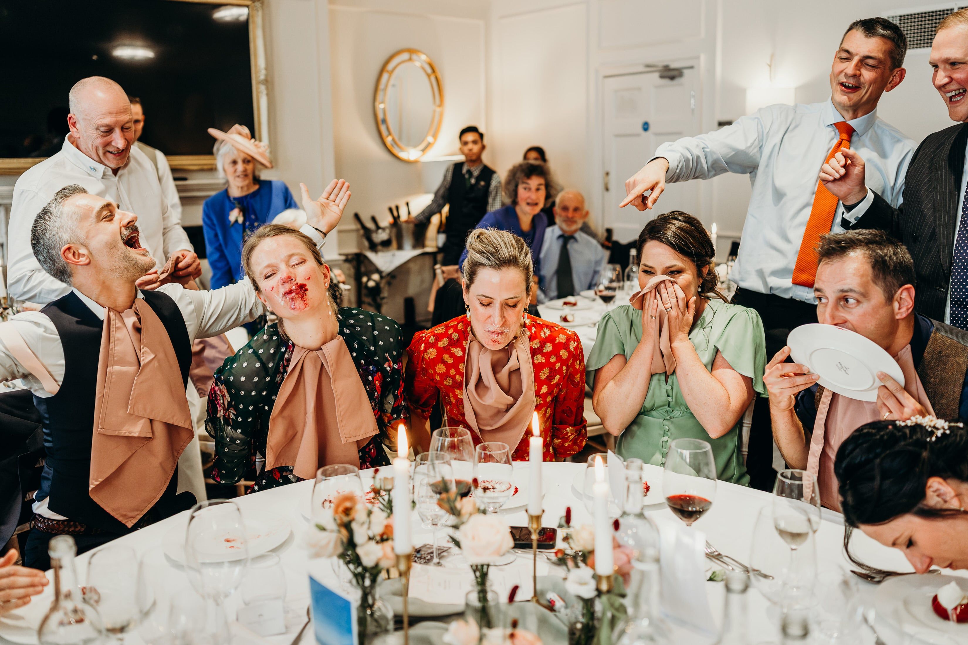 Guests at a wedding reception table are mid-action during a game, with several covered in cake or